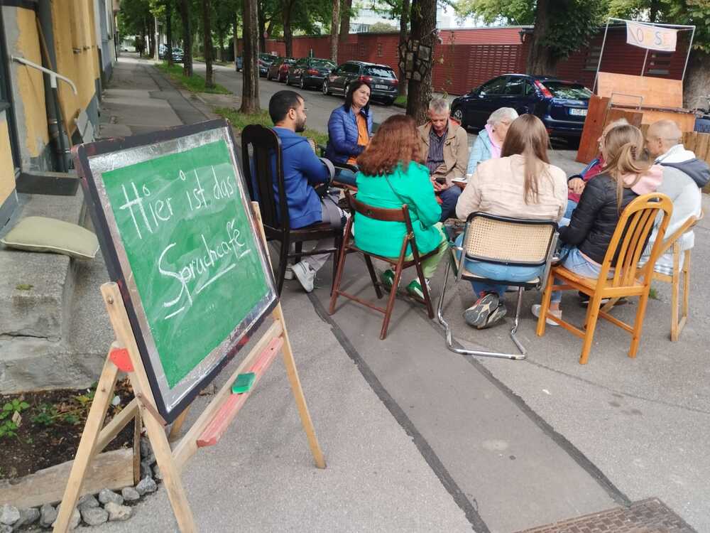 Menschen sitzen im Stuhlkreis vor dem Quartierszentrum, mit einer Tafel im Vordergrund auf der „Sprachcafé